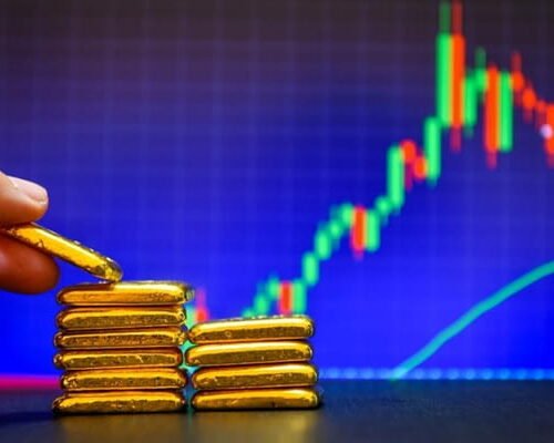 Hand placing gold bars on a stack with a rising financial market chart in the background.